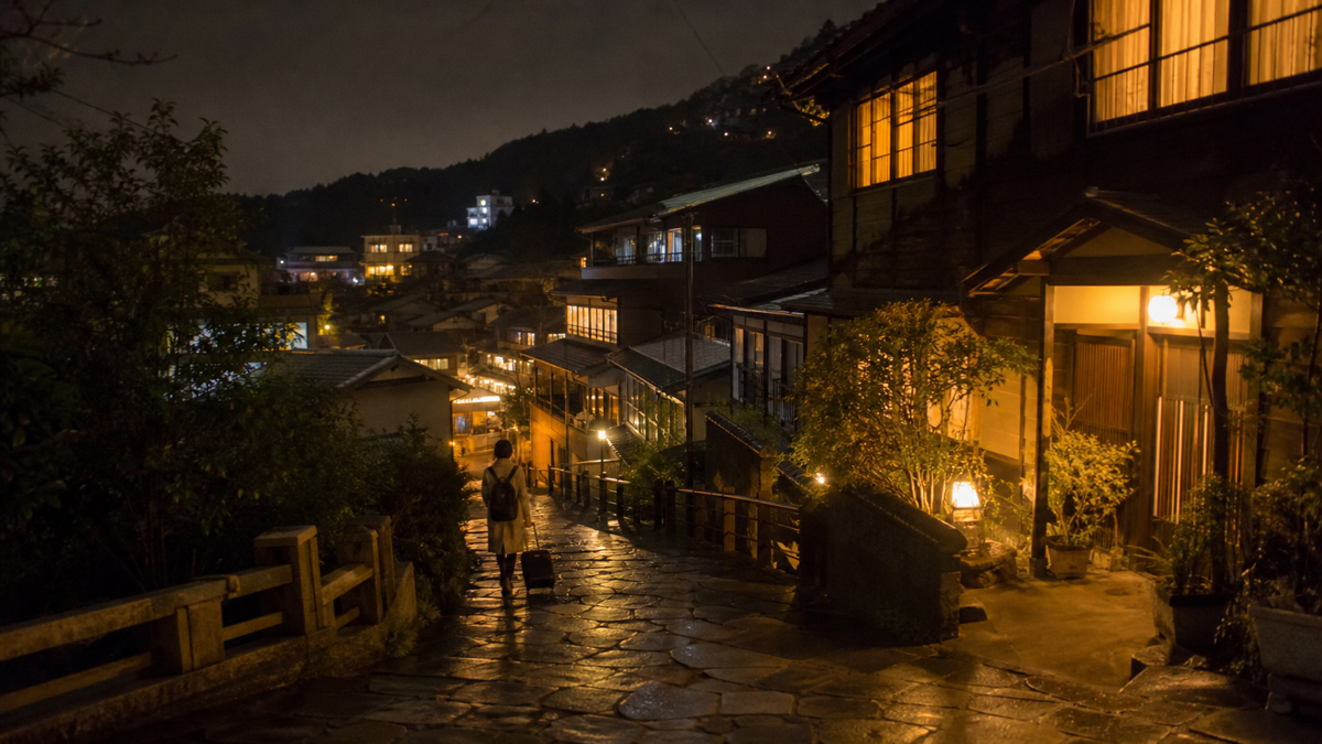 夜の山あいの温泉街を小さな旅行バッグで歩くイメージ